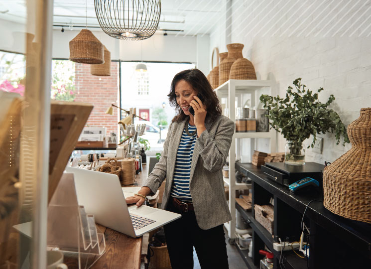 Woman managing small business finances on phone and laptop in retail store.