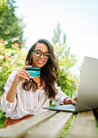 Young woman using a laptop and holding a credit card while sitting outside on a sunny day.