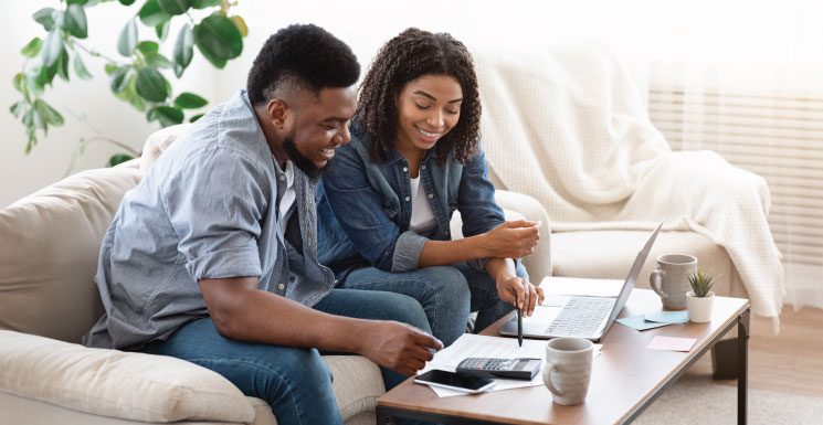 A smiling couple sits on a couch reviewing finances together on the table in a bright living room.