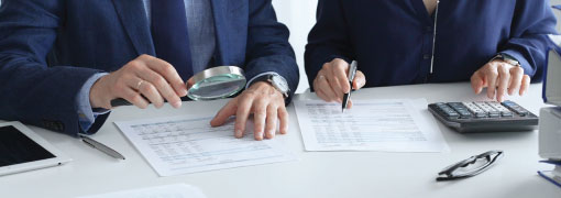 Two professionals reviewing documents closely with a magnifying glass and calculator.