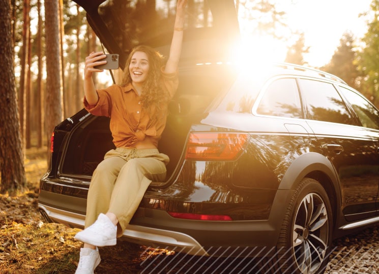 Person taking selfie while sitting on car trunk in sunny outdoor setting