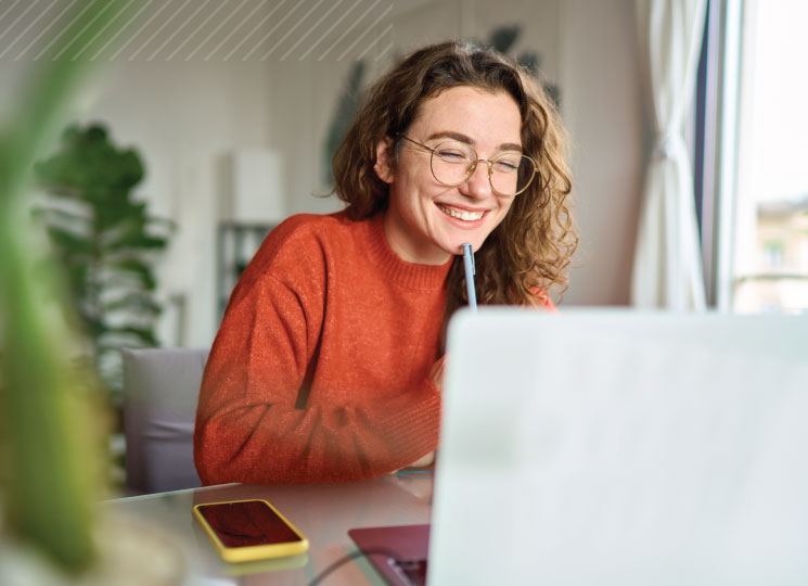 Woman smiles while using a laptop at a table near a window.