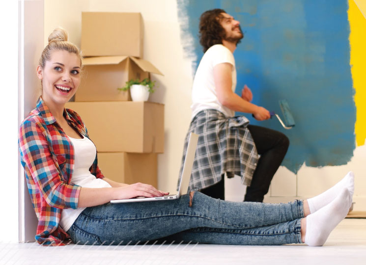 Woman sits on the floor using a laptop in a new home with moving boxes while a man paints a wall in the background.