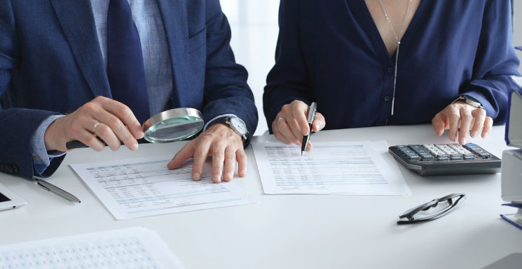 Two professionals reviewing documents closely with a magnifying glass and calculator.