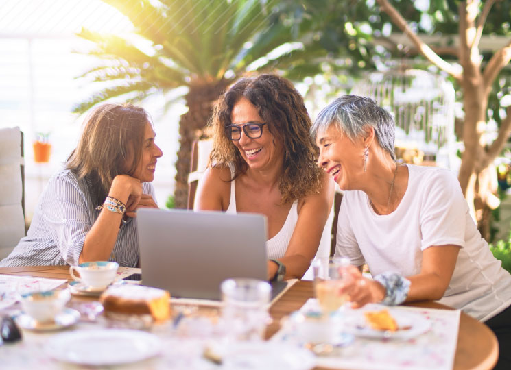 Three women laugh together while looking at a laptop at an outdoor café.