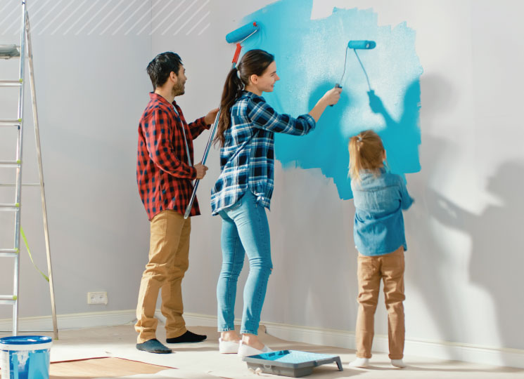 Man, woman, and child paint an interior wall with rollers, with a ladder and paint tray nearby.