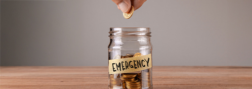 Hand placing coin in jar labeled EMERGENCY showing what is an emergency fund for savings.