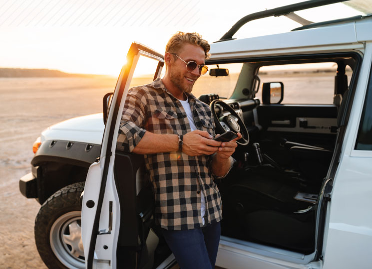 Man checking his phone while leaning against an open Jeep at sunset.