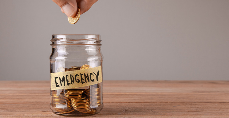Hand placing coin in jar labeled EMERGENCY showing what is an emergency fund for savings.