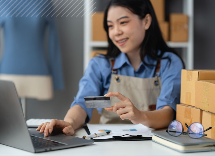 Small business owner works at a desk on a laptop while holding a credit card, with shipping boxes stacked nearby.