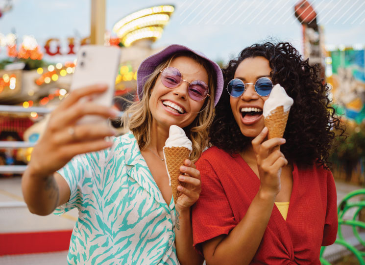 Two friends take a selfie while holding ice cream cones at an amusement park.