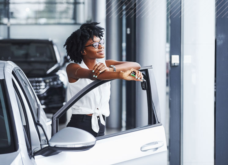 Woman holding car keys while leaning on an open vehicle door at a dealership.