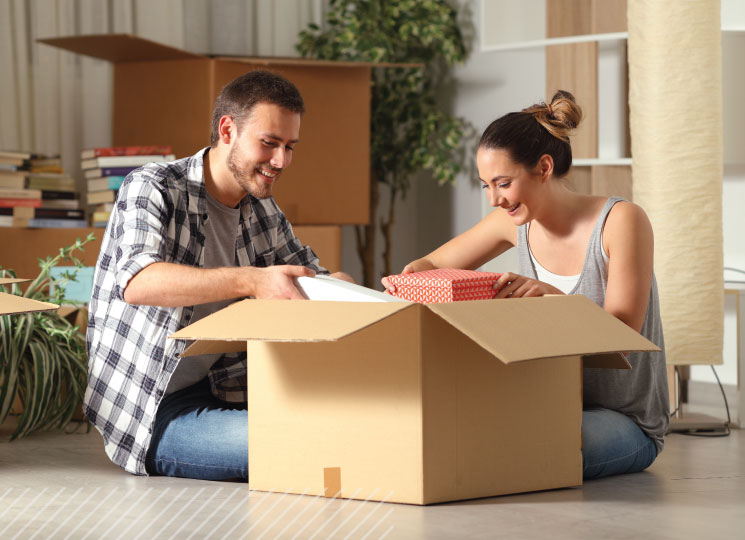 Couple sits on the floor unpacking household items from a large moving box in their living room.