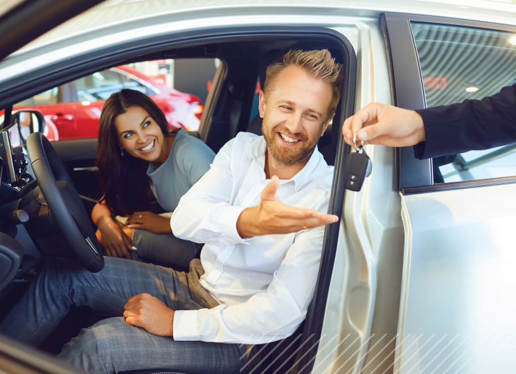 Smiling man seated in a car reaches for keys while a woman sits in the passenger seat.